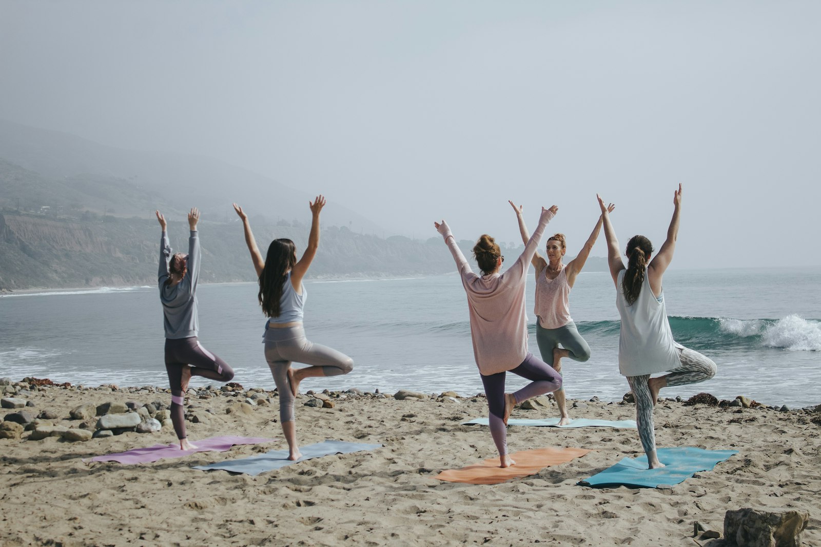 Women practicing yoga tree pose on beach mats at soft daylight shoreline