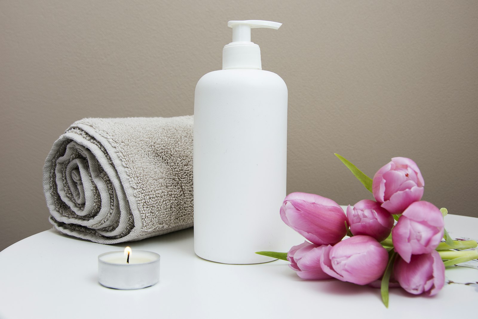Unbranded white pump bottle with rolled towel, pink tulips, and tea light — spa amenity daylight